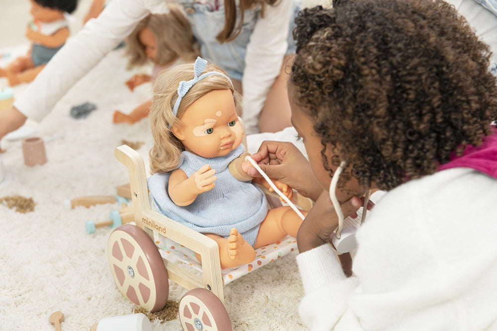 a little girl playing doctor with her Miniland Educational Vitiligo doll in their new wheelchair for dolls