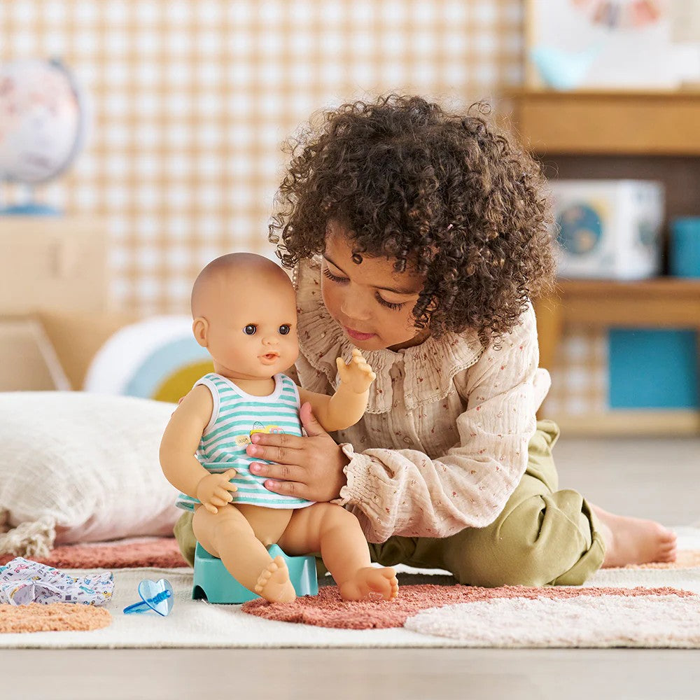 Young girl pressing Corolle Baby Paul drink and wet doll belly while doll sits on teal potty chair during pretend potty training play