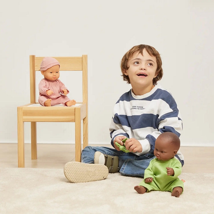 Child with Down Syndrome sitting on the floor with two down syndrome dolls, one on a chair, against a plain background