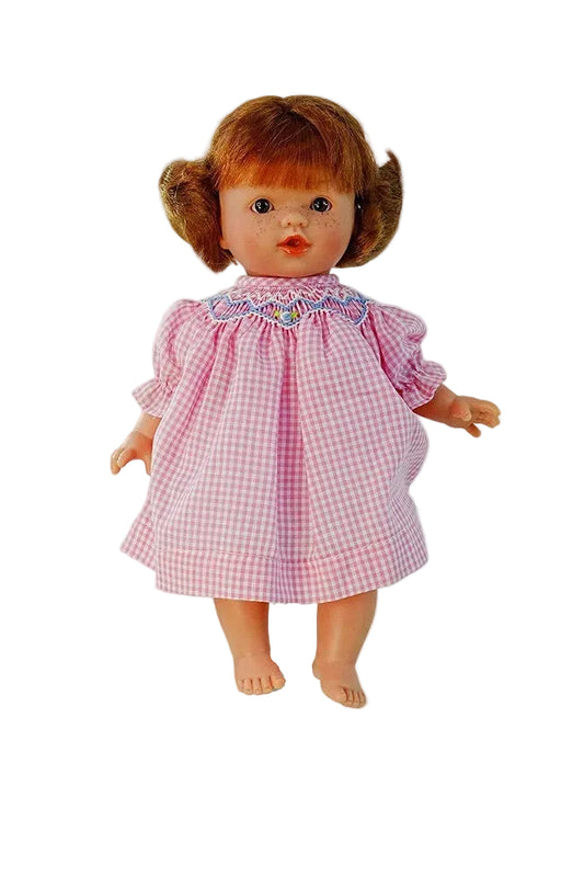 a small lifelike redhead doll with brown eyes dressed in a pink gingham dress with a smocked and embroidered colar