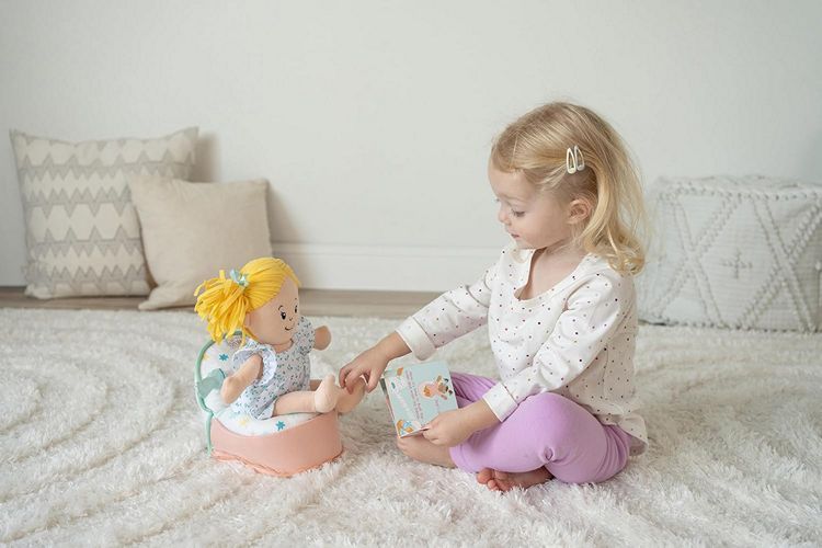 Young toddler girl reading My New Potty board book to Baby Stella doll seated on soft plush potty during pretend potty training play